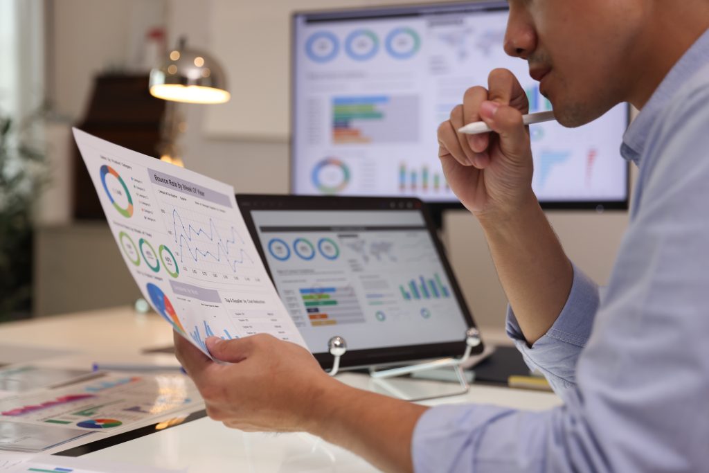 Person reviewing printed charts and graphs at a desk with a laptop and monitor displaying data visualizations in the background.