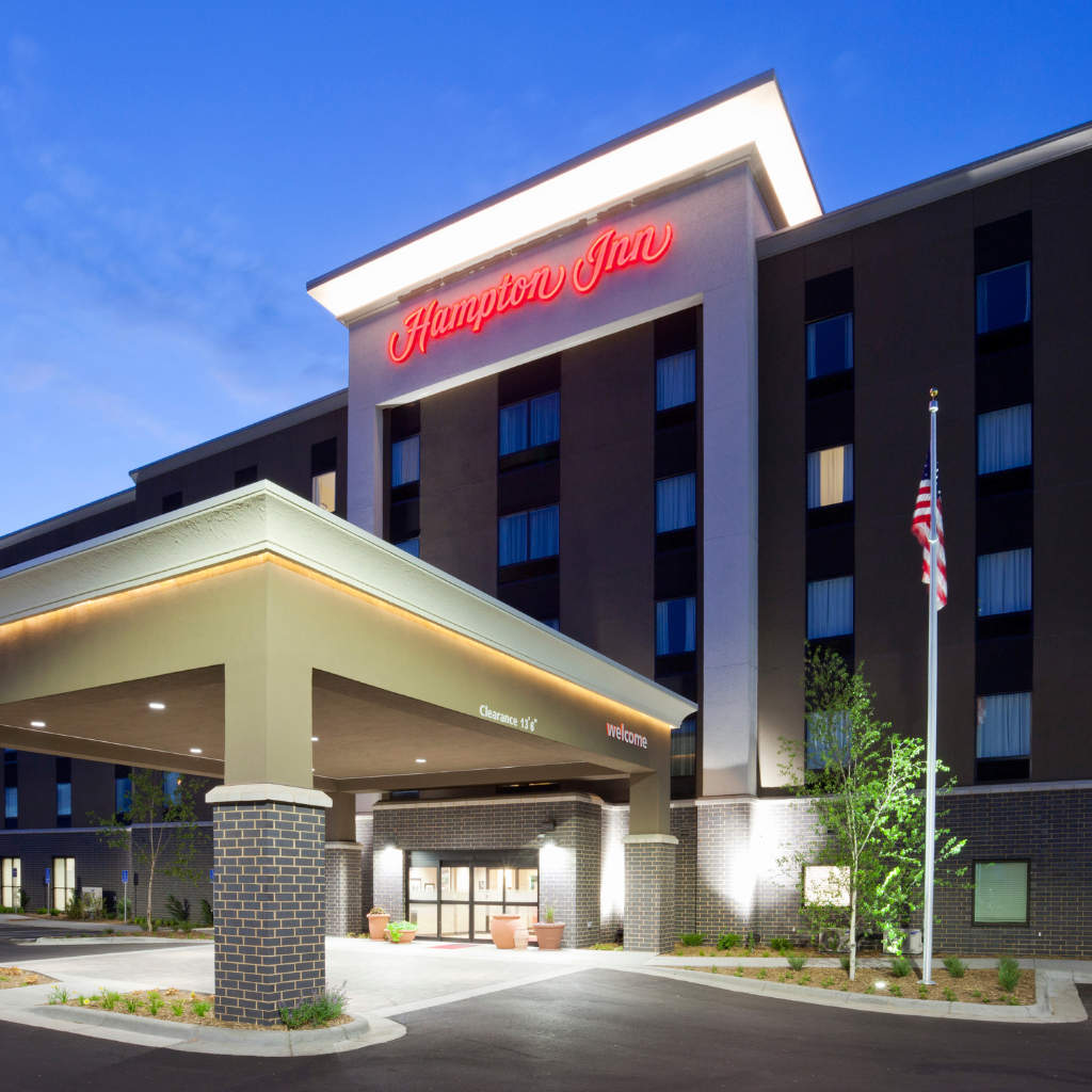 Exterior of a Hampton Inn hotel at dusk with a covered entrance, brick columns, and an American flag nearby.