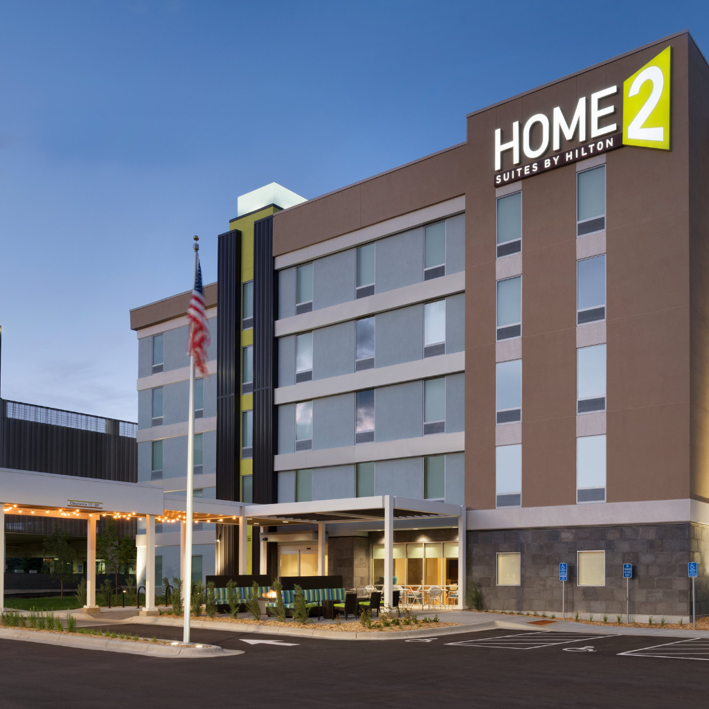 Exterior view of a Home2 Suites by Hilton hotel with a covered entrance and outdoor seating under a blue sky.