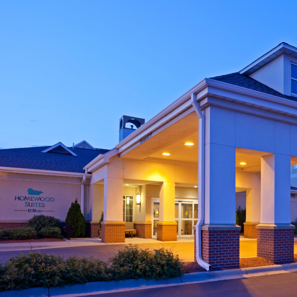 Exterior of a Homewood Suites by Hilton hotel at dusk, showing a covered entry with lit lamps and brick columns.