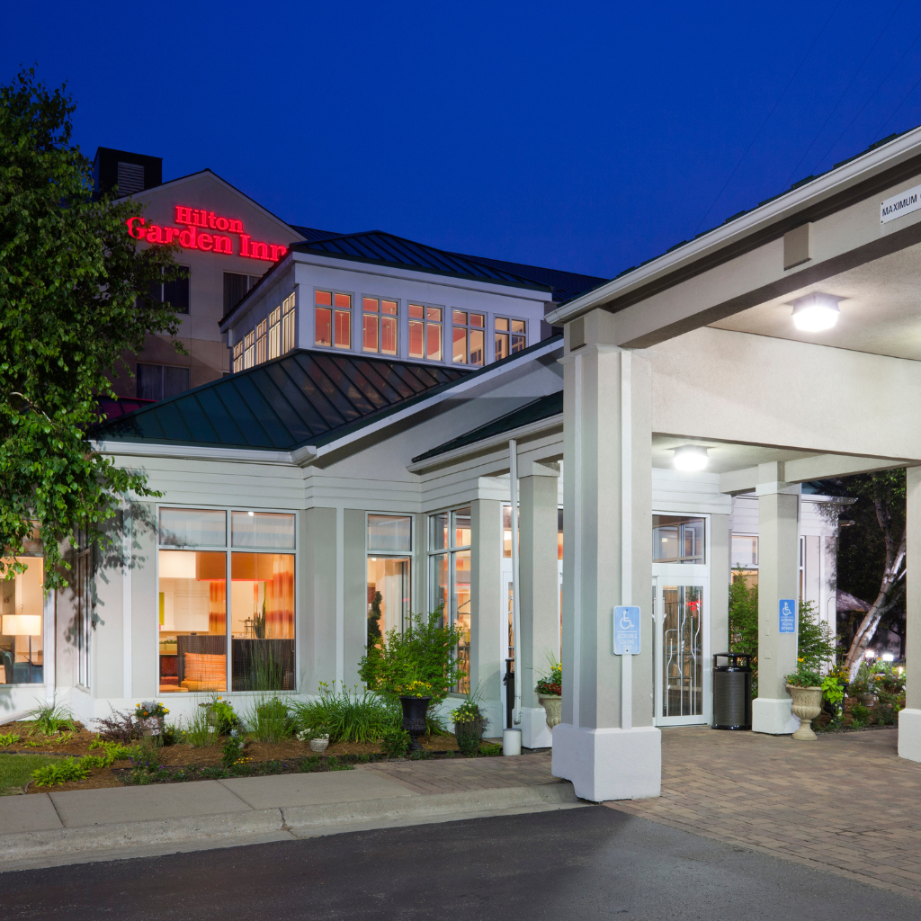 Hotel entrance of Hilton Garden Inn at dusk with lit windows and a covered drop-off area, handicap parking signs visible