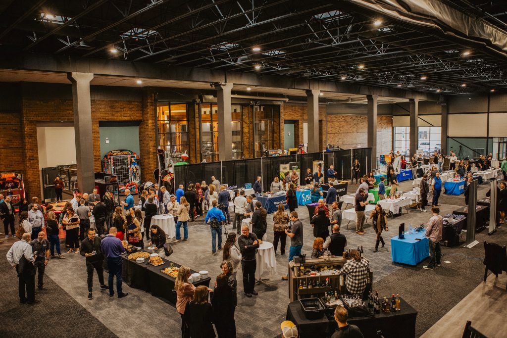 Busy indoor expo hall with numerous vendor booths, attendees browsing and mingling around catering tables.